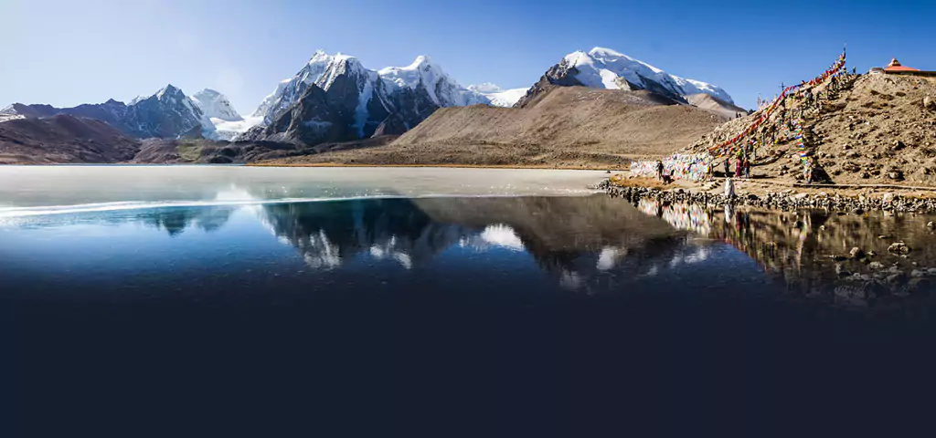 Serene lake with snow-covered mountains and prayer flags, a picturesque spot offered by tour operators in Kolkata.
