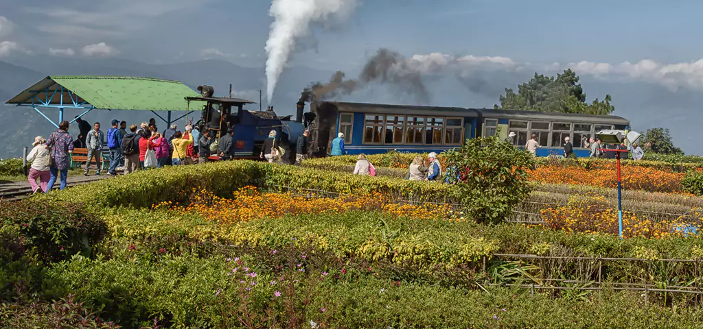Steam train at station with flower gardens and mountain backdrop, a scenic trip recommended by travel company in kolkata.